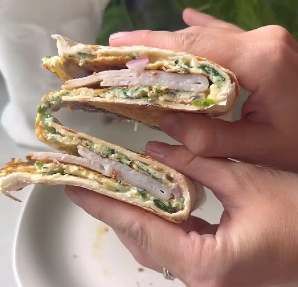 Hand holding a crispy egg tortilla wrap stuffed with turkey, spinach, and eggs on a rustic table.